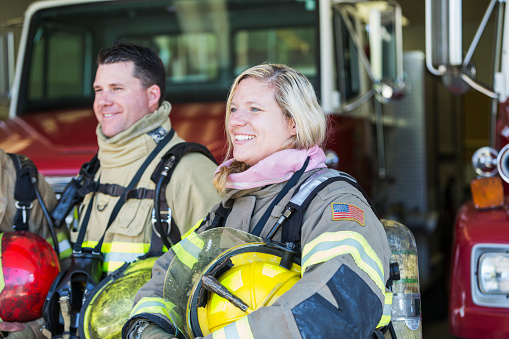 A female firefighter standing with a male colleague in front of a fire truck, wearing protective gear, holding their helmets.  She has a patch of an American flag on her arm.