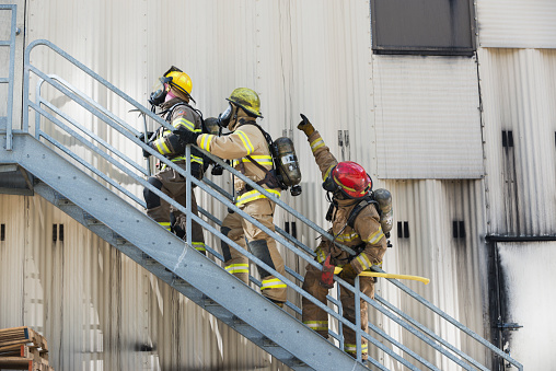 Firemen on telescopic ladder extinguishing strong fire with from a fire fighting firehose nozzle in rescue operation.Thick grey smoke and fire from the windows without glass of industrial warehouse.