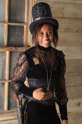 Cute black girl wearing a stovepipe hat and a Victorian-style dress, posing with a revolver and gunbelt - Steampunk fashion.