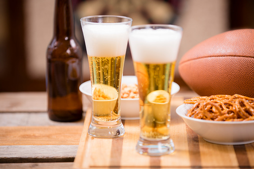 Sports bar, pub setting.  An American football, pretzels, peanuts, beer in glasses and in a bottle are all on the wooden bar counter top.  A dartboard is in the background.  championship game party!   