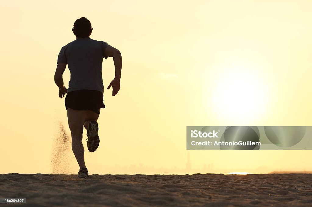 Back view silhouette of a runner man running Back view silhouette of a runner man running on the beach at sunset with sun in the background Men Stock Photo