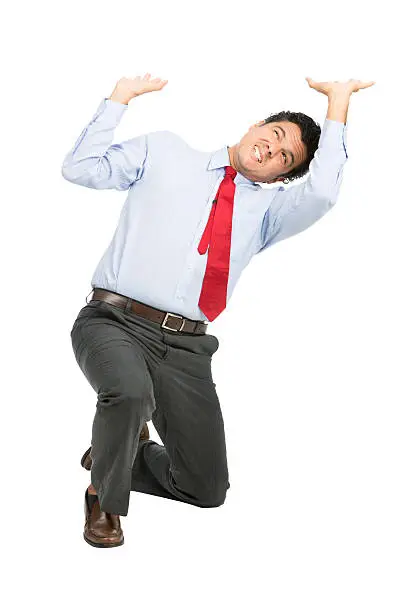 A stressed latino businessman in business clothes on knee using arms pushing up, resisting against crushing imaginary weight, object under heavy stress, feeling pressure. Isolated on white background