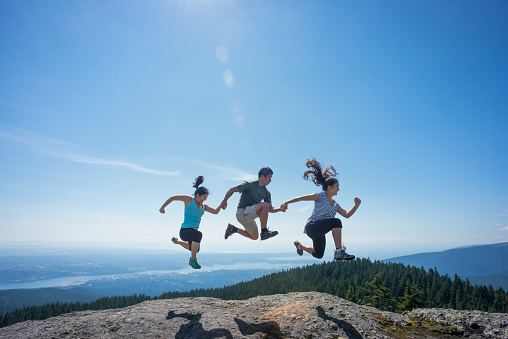 Mature Asian father and mixed-ethnicity teenage daughters hold hands while jumping on the top of a mountain, at the edge of a cliff.  Wide angle view of forest and City of Vancouver below and in the background.  Mount Seymour Provincial Park, North Vancouver, British Columbia, Canada.  Lens flare.