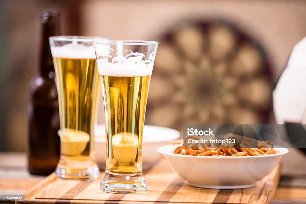 Pub food. Snacks, beer, soccer ball. Sports bar atmosphere. Sports bar, pub setting with a soccer ball, pretzels, beer in glasses and a bottle on a wooden bar counter top.  A dartboard is in the background.  No people in image.   2015 Stock Photo