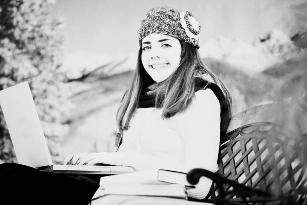 Young Latin descent woman uses laptop computer outside during the winter season.  A stack of books is beside her on the park bench.  Winter mountains in the background.  College campus or park setting.  Winter clothing.  Black and white image with grain added for effect. 