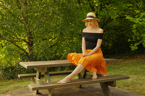 Photo of a young blond woman sitting on a bench in a park with trees in the background.  Young woman is wearing orange skirt, black off-the-shoulder top and hat. Taken on a Pentax K-3 at 1/160, f-5 and ISO200.