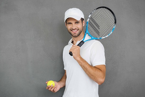 Happy young man in sports clothes carrying tennis racket on his shoulder and smiling while standing against grey background
