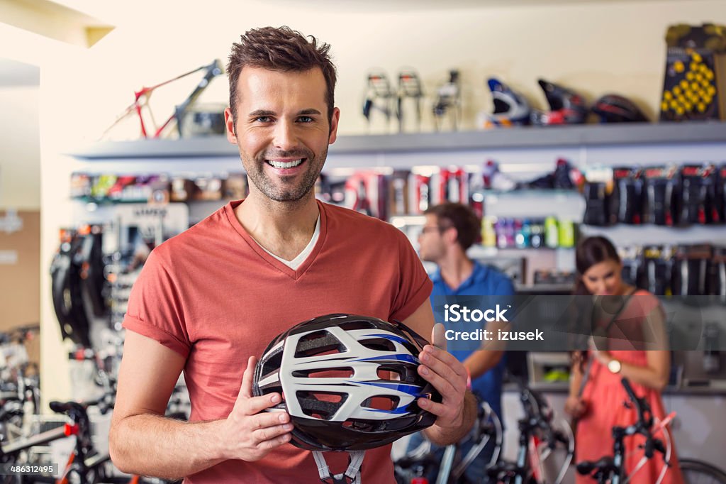 Man buying a helmet Bike store: man holding a bicycle helmet and smiling at camera with two people in the background. Looking At Camera Stock Photo