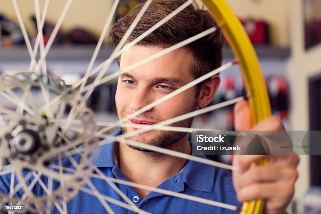 Young man with a bicycle wheel Portrait of young sales clerk in a bike store looking at bicycle wheel. 20-24 Years Stock Photo