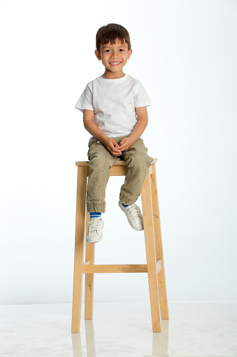 Cute young boy sitting on a stool against a white background, smiling for the camera.