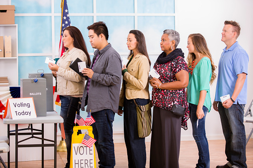 Multi-ethnic, mixed age group of USA citizens stand in line to cast their ballot in the November US elections at a local polling station. A table with ballot boxes, and \