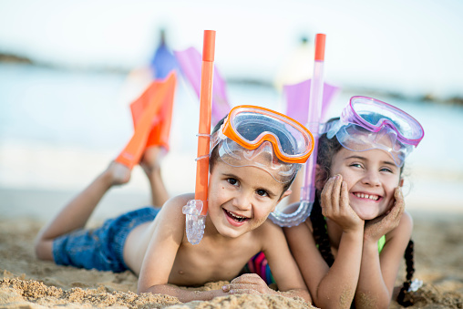 A brother and sister are on vacation with their family in Hawaii, they have their snorkeling gear on and are ready to get in the water - they are both smiling and looking at the camera.