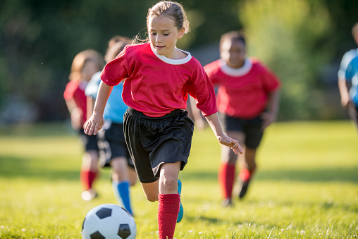 A little girl is kicking the ball up the soccer field on a beautiful sunny day as the rest of her teammates are trying to keep up.