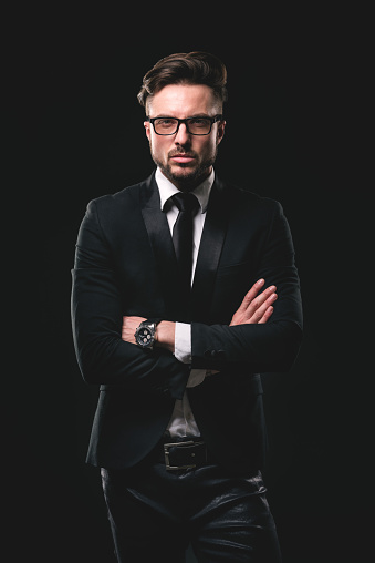Studio portrait of young man with glasses wearing necktie and black suite. He is also wearing black leather pants and holding arms crossed. Black background.
