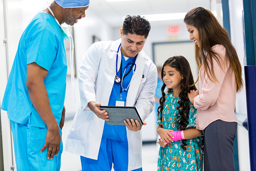 Senior adult African American surgeon and mid adult Hispanic male doctor are talking to pediatric patient and her mother in hallway of hospital. Doctor is using digital tablet to show patient her chart or results. Patient is elementary age Hispanic girl, who is wearing a hospital gown and a cast on her wrist.