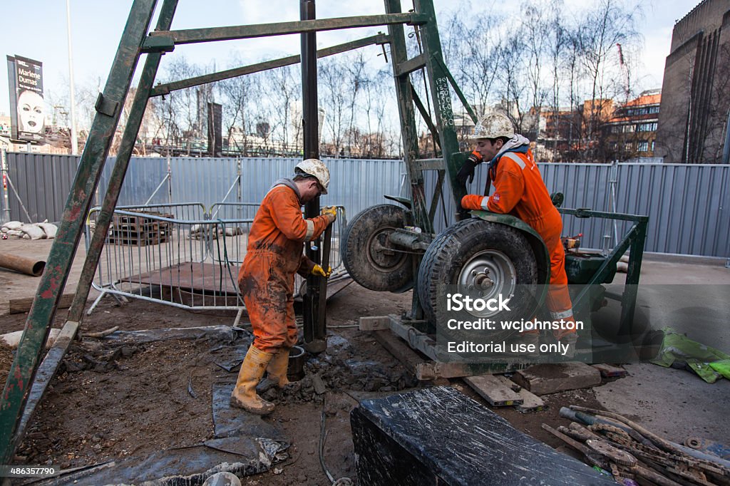 borehole drilling rig London, England - March 10, 2015: Workmen in orange overalls and white hardhats outside. they have yellow gloves and boots and are next to a borehole drilling rig. a fence can be seen. One man is cleaning out the clay from the dril bit and the other holds it steady. Behind is the London skyline. 2015 Stock Photo