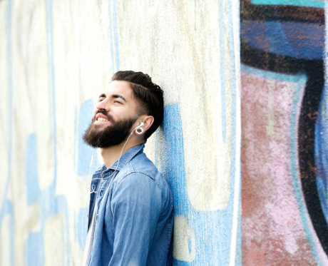 Close up portrait of a young man with beard smiling and listening to music on earphones outdoors