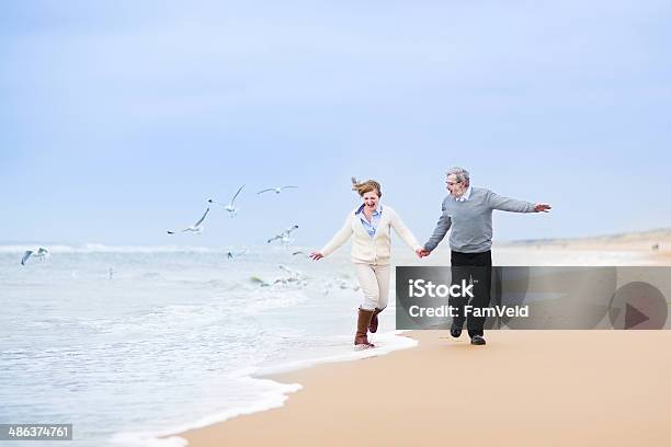 Happy Mature Couple Running At Beautiful Winter Beach With Seagulls Stock Photo - Download Image Now