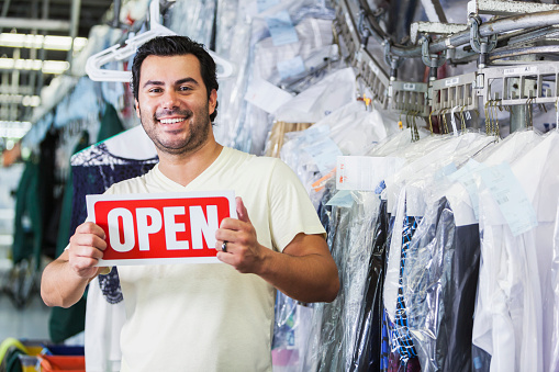 An Hispanic man working in a dry cleaning store, standing in front of racks of drycleaned clothing hanging on hangers, holding an OPEN sign and smiling at the camera.  He is wearing a white t-shirt and blue jeans.