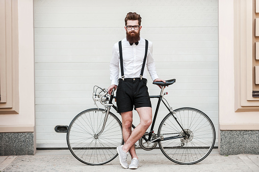 Full length of handsome young bearded man leaning at his bicycle and looking at camera while standing outdoors