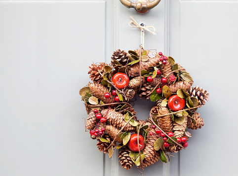 Seasonal wreath for Autumn and Christmas decorated with pine cones, red apples and red berries, bound with a vine, hanging on a grey painted, panelled door. Good copy space.
