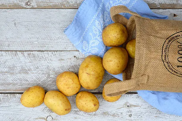 Organic fresh uncooked potatoes in a jute bag on a old weathered wooden white shelves background with vintage blue kitchen tea towel with kopie space