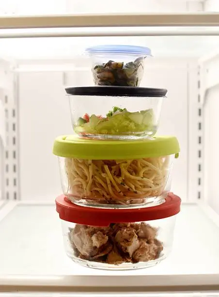 This image is of stacked food storage containers in the fridge filled with leftovers of diced chicken, spaghetti, diced cucumbers and diced mushroom.  Each food storage container has a different color lid. 