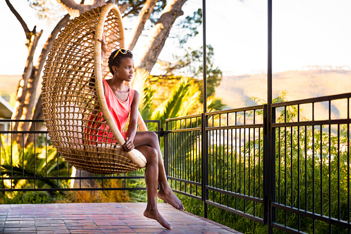 Young african woman sitting in a hanging chair in patio looking away