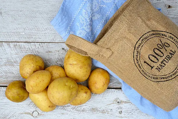 Organic fresh uncooked potatoes in a jute bag on a old weathered wooden white shelves background with vintage blue kitchen tea towel with kopie space