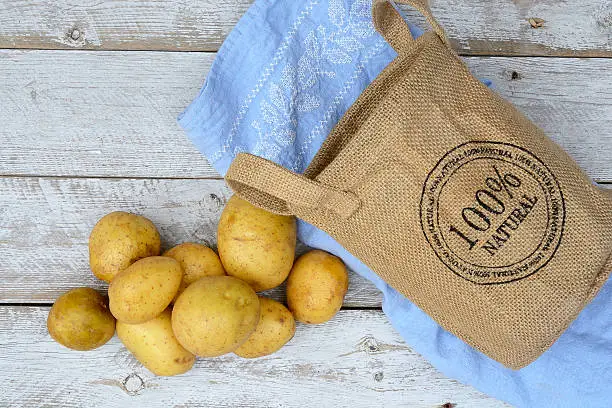 Organic fresh uncooked potatoes in a jute bag on a old weathered wooden white shelves background with vintage blue kitchen tea towel with kopie space