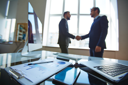Business objects at workplace with businessmen handshaking on background