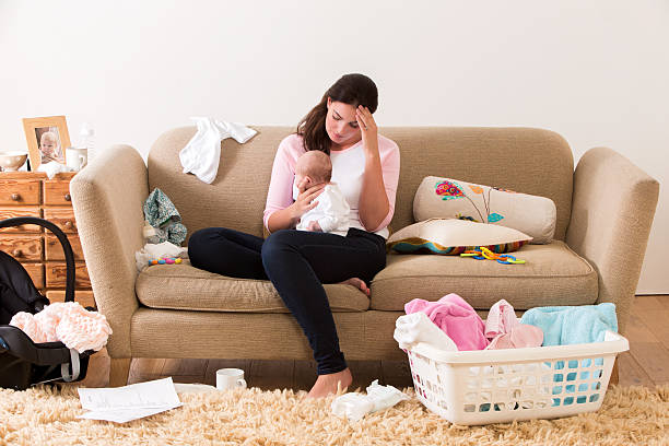 Stressed Mother with Baby Mother with her baby on her knee. Her head is lowered as she sits on a sofa looking tired and stressed. The room looks untidy with clothes, nappies and other items scattered around the room. postpartum depression stock pictures, royalty-free photos & images