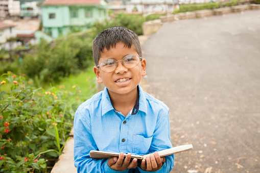 Cute boy living in northeastern India is excited to go back to school. The child is holding a book and wearing eyeglasses. He is outdoors by alongside a rural road near his village, which is seen in the background.  Elementary-aged student.