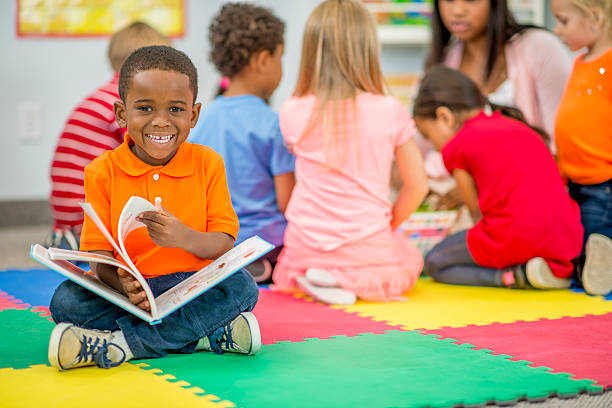 Little Boy Looking at Picture Book in Preschool A little boy in preschool is sitting on a foam mat with her classmates and is holding a picture book - he is smiling and looking at the camera. boy and girl reading stock pictures, royalty-free photos & images