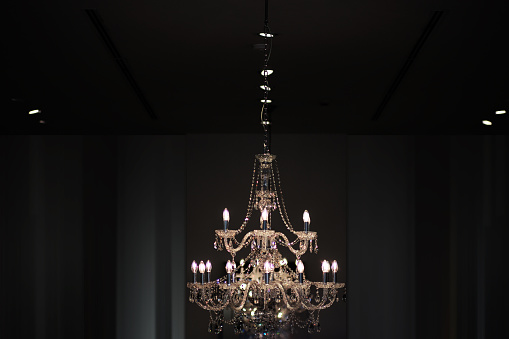 Closeup of one big beautiful old electric chandelier with candles and lots of decorative crystal beads hanging on ceiling and brightly lit on dark indoor background, horizontal picture