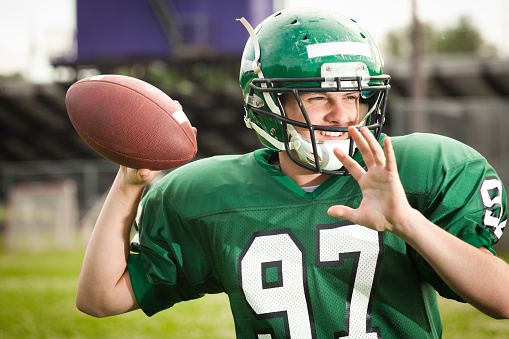 American football player quarterback in a game. A college American football player holding a football in his hand ready for throwing a pass to a receiver. He is wearing a green football jersey and helmet. Photographed in the stadium in vertical format.