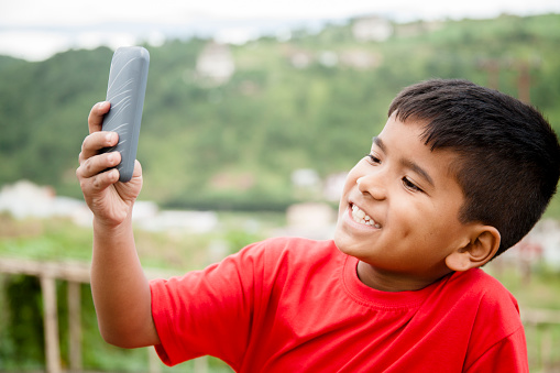 Elementary age, Indian or Latin descent boy using a smart phone outdoors.  He is using social media, texting a friend, video chatting, or learning about his world on the internet. He enjoys exploring new things. He wears a red shirt and a smile. He is sitting alongside a rural road. His village can be seen behind him.  Rural mobility.