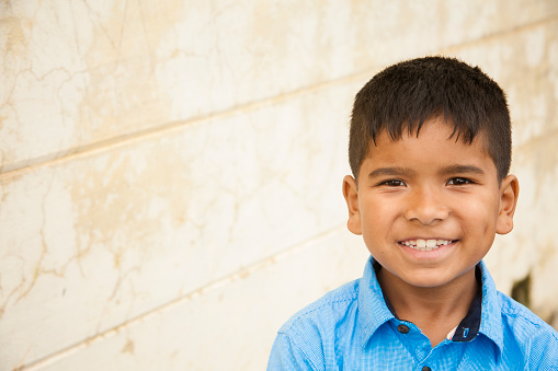 Headshot of cute little Latin or Asian descent boy outdoors in front of a tan colored, textured wall. The elementary age boy wears a big smile and a blue collared shirt. Copyspace to left.  Portrait, close-up style image.