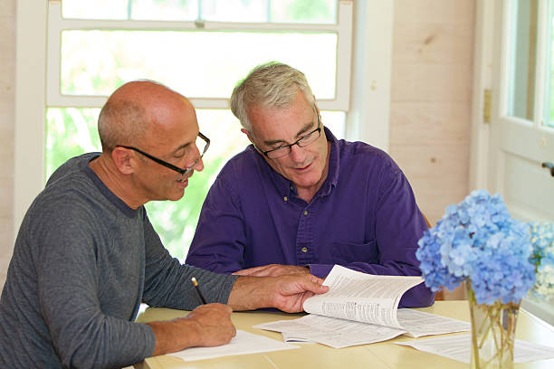 Senior Gay Male Couple Working Together on Financial Documents Older, mature, senior gay male couple, affectionate and in love, working together on financial documents at their dining room table at home. homosexual couple stock pictures, royalty-free photos & images