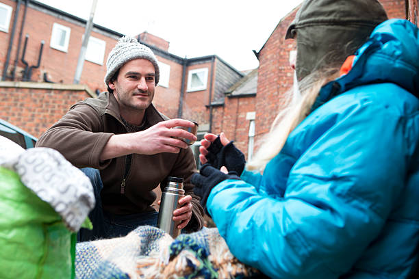 Homeless Woman Receiving Help Homeless woman sitting on the street in the cold. A kind male offers her a hot drink from a flask. beggar stock pictures, royalty-free photos & images