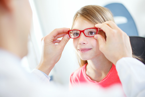 Little brown eyed  girl visiting optician.She's having appointment for testing new glasses.Middle aged male optician is gently adjusting new frame and glasses on girl's face.