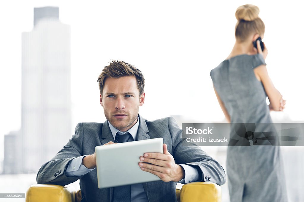 Businessman with digital tablet Businessman sitting in a chair and using a digital tablet with a businesswoman talking on phone in the background. 20-24 Years Stock Photo