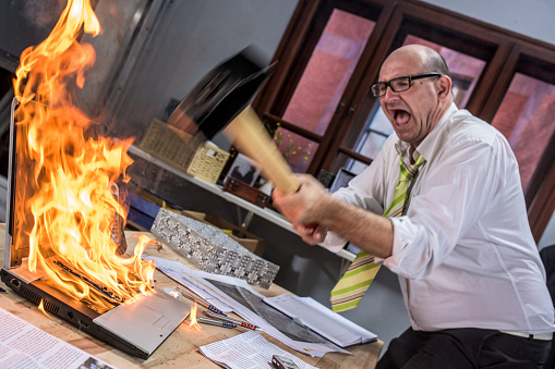 Mature adult businessman smashing laptop on fire with hammer