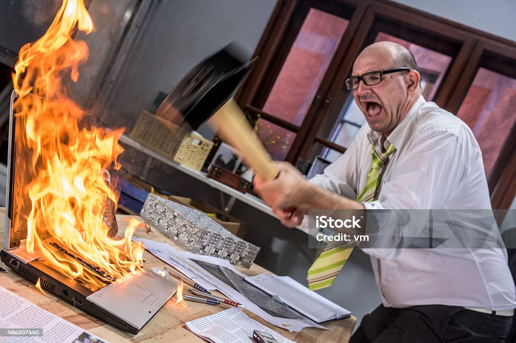 Mature adult businessman smashing laptop on fire with hammer Frustrated businessman hitting laptop on fire with hammer. Computer Stock Photo