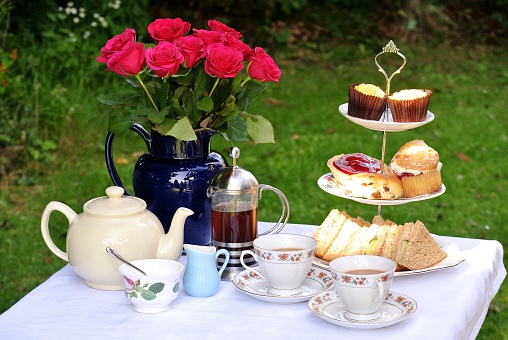 Photo of a table set for afternoon tea for two. There is a three-tiered cake stand containing sandwiches, cream cakes, a scone, a strawberry tart and cupcakes. There is also a tea pot, a cafetiere of coffee and two china cups and saucers. The table is decorated with a white table cloth and a large, blue jug containing a bunch of pink roses.