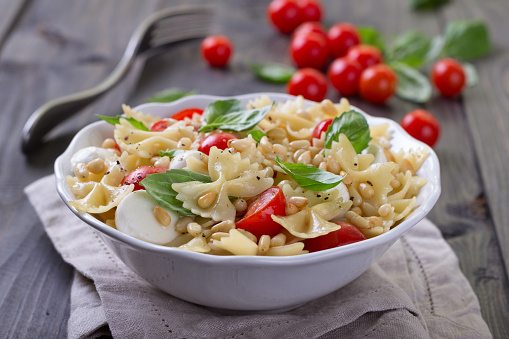 Pasta salad with tomato, mozzarella, pine nuts and basil in a white ceramic bowl on a wooden table