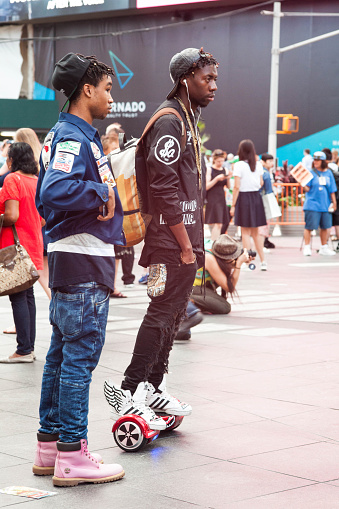 New York City, NY, USA - August 27, 2015: Two stylish, African American young men chill in Times Square. Both with reverse caps. Guy in right with trendy red Sprayground Hoverboard / skateboard, ripped jeans, gold chains & Winged Adidas sneakers. His friend with SCCA, Amateur road racing, Central Florida Jacket and pink Timberland's.