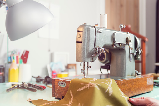 Portrait of a dressmaker's cluttered studio space showing with a retro styled, electric sewing machine sitting on a desk surrounded by the associated paraphernalia of dressmaking. Very shallow focus point on the machine's needle, colour, horizontal, with a high key light effect, giving a nice out of focus background.