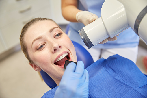 people, medicine, stomatology, technology and health care concept - close up of dentist putting intraoral shield to female patient mouth and assistant directing x-ray machine at dental clinic