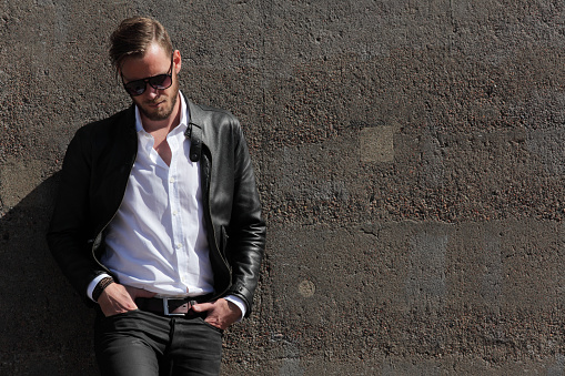 A man in his 20s wearing a black leather jacket and white shirt, leaning back against a wall of stone on a sunny summer day, 